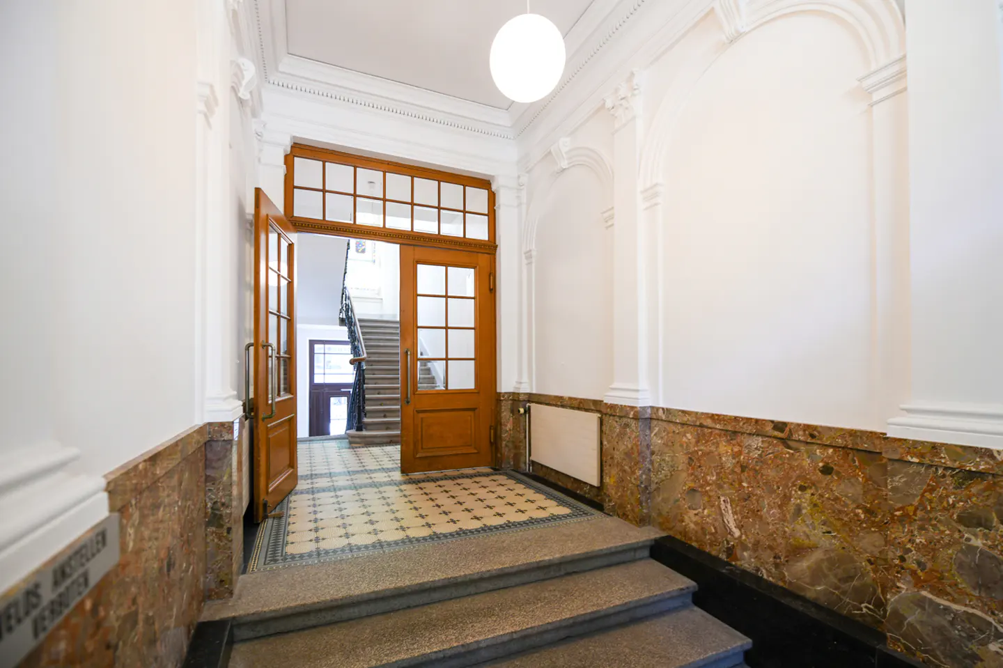 Interior view of a building entrance with marble walls, a wooden door, and stairs leading to a tiled floor. A staircase is visible through the open door.