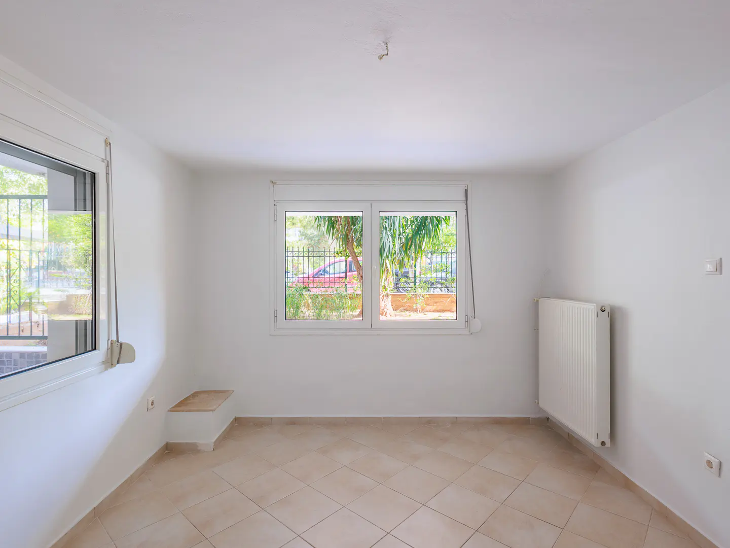 Bright, empty room with white walls, beige tile floor, and two windows. A radiator is on the right wall. Outside, trees and a car are visible.