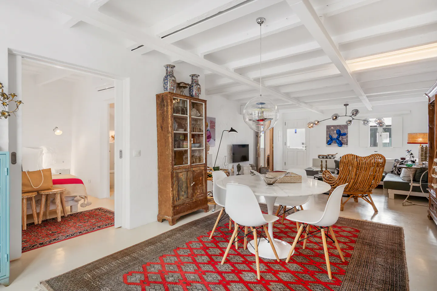 Bright, open-concept living space with white walls, exposed beams, and a red patterned rug. A round table with white chairs sits near a rustic wooden cabinet.