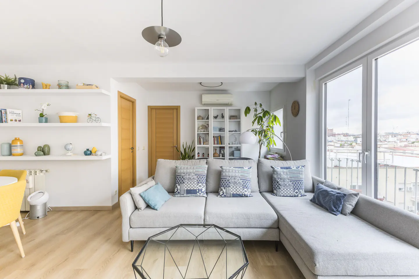 Bright living room with a gray sectional sofa, blue pillows, and a geometric glass coffee table. Shelves and a large window with a city view.