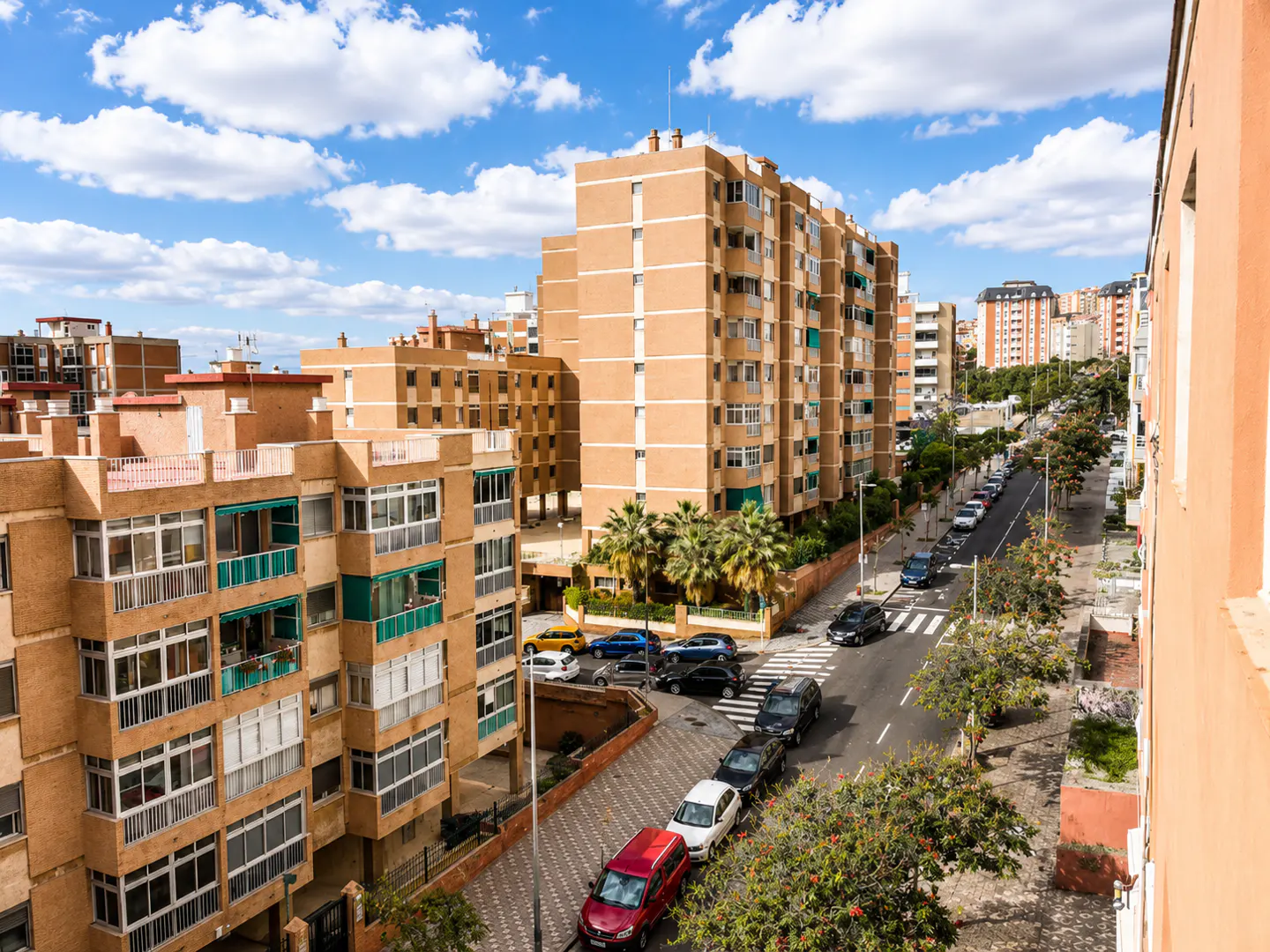 View of a street lined with peach-colored apartment buildings under a blue sky with white clouds. Cars are parked along the street.