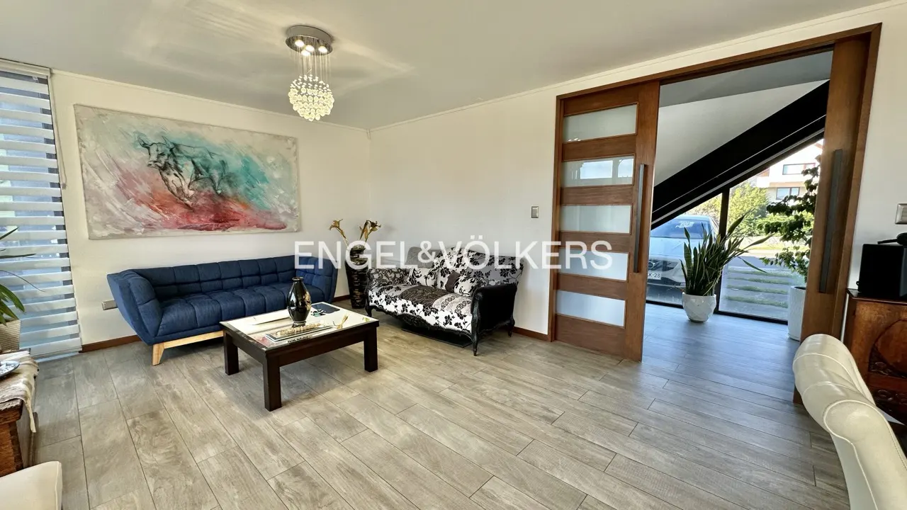 A bright living room with a blue sofa, patterned loveseat, and a dark wood coffee table. A large painting hangs above the sofa. A sliding glass door leads to the outside.