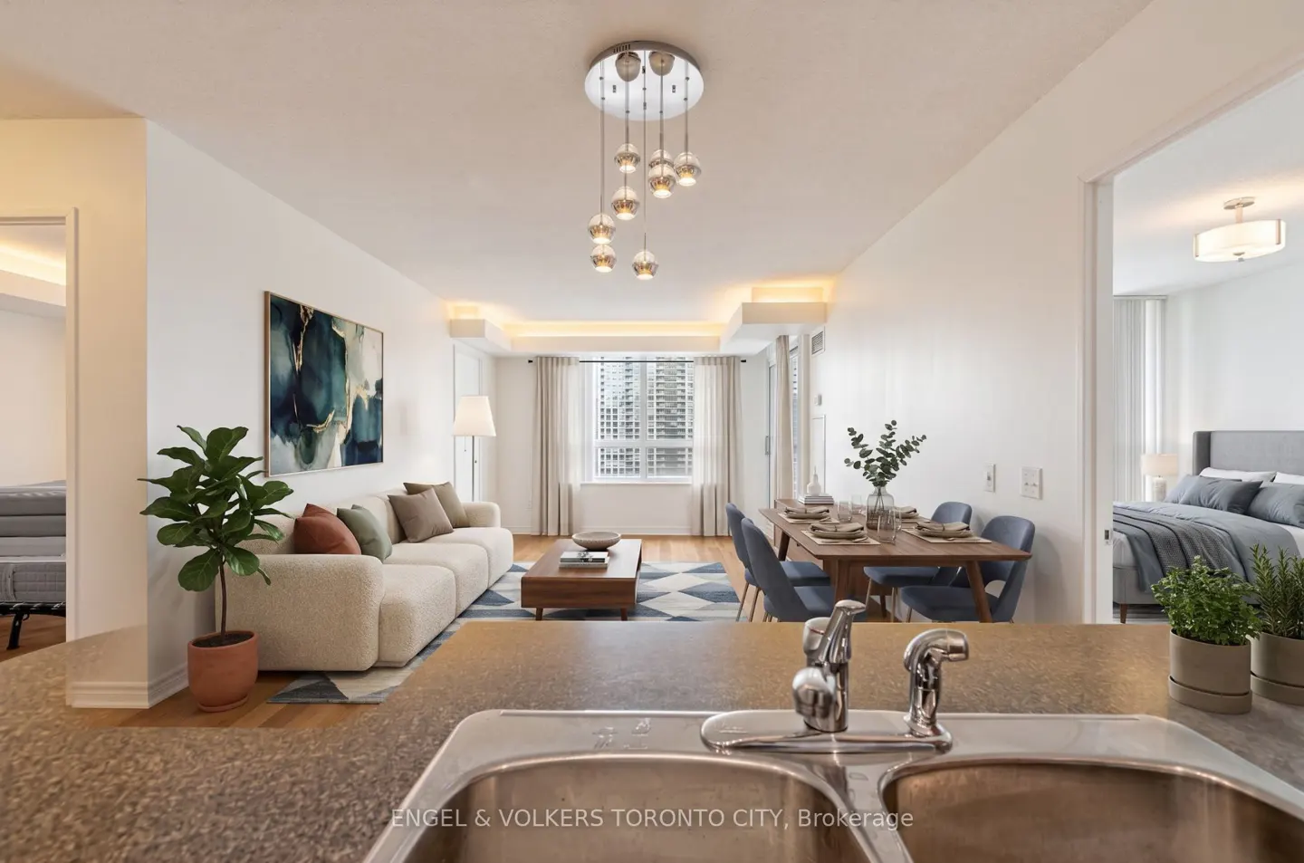 A bright, modern condo interior with a kitchen sink in the foreground, a living room with a white sofa, and a dining area with a wooden table.
