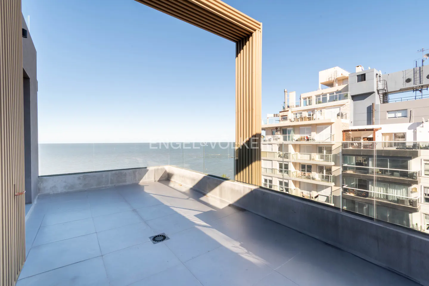 Balcony view with ocean and city buildings. The balcony has light gray tiles and a wooden frame. The sky is blue.