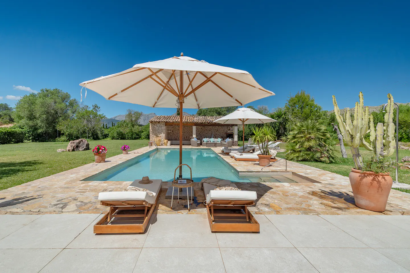 Outdoor pool area with two lounge chairs, white umbrellas, and a stone building in the background under a clear blue sky.