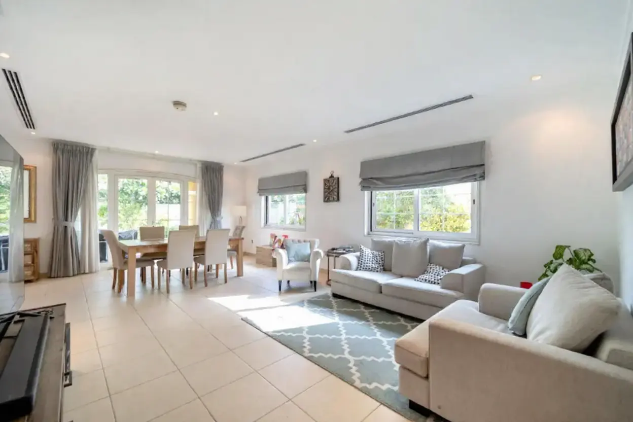 Bright, open-plan living and dining area with neutral decor. A light wood dining table and gray sofas sit on tiled floors with a patterned rug.