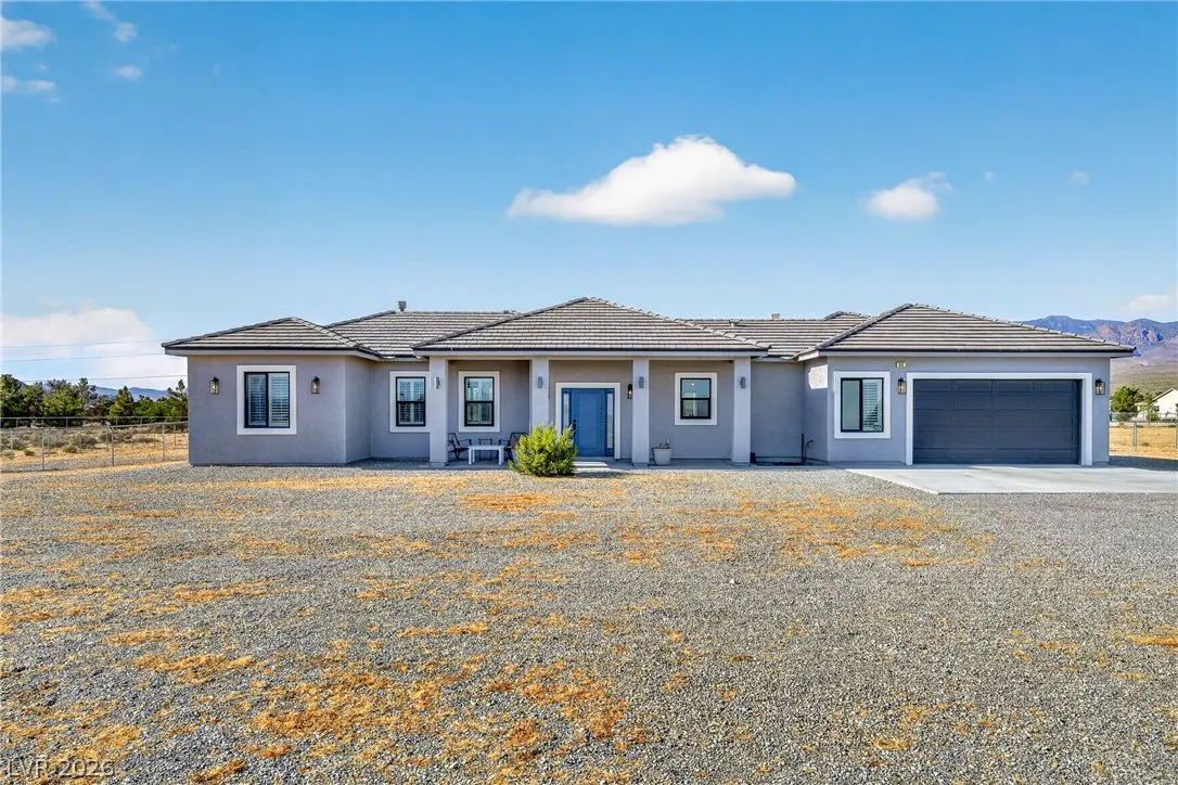 Single-story gray stucco home with a blue front door, tile roof, and attached garage on a large gravel lot under blue sky.