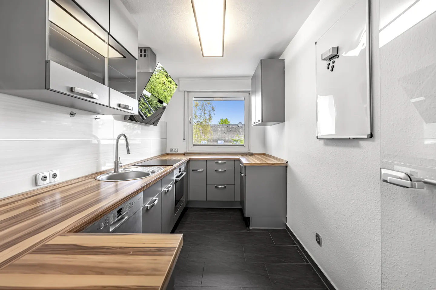 A modern kitchen with gray cabinets, wood countertops, and black tile flooring. A window provides natural light, and a whiteboard hangs on the wall.