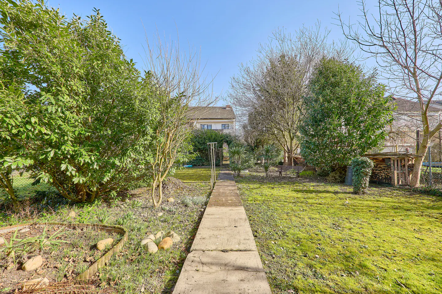 A long concrete path leads through a green lawn and trees to a house under a blue sky.