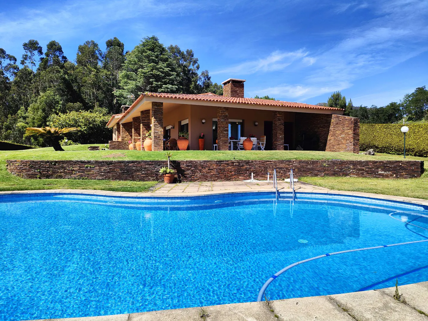 A single-story house with a red tile roof overlooks a blue swimming pool on a sunny day. Trees are in the background.