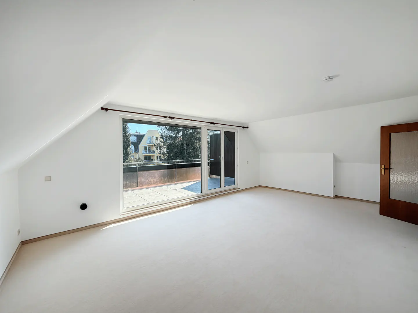 Bright, empty room with white walls, light carpet, and a sliding glass door to a balcony. A brown door is on the right.
