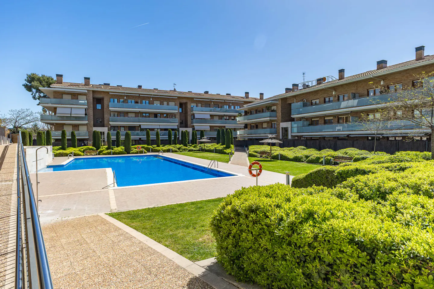 Apartment complex with a blue swimming pool, green grass, and trimmed bushes on a sunny day. A red life preserver is visible.