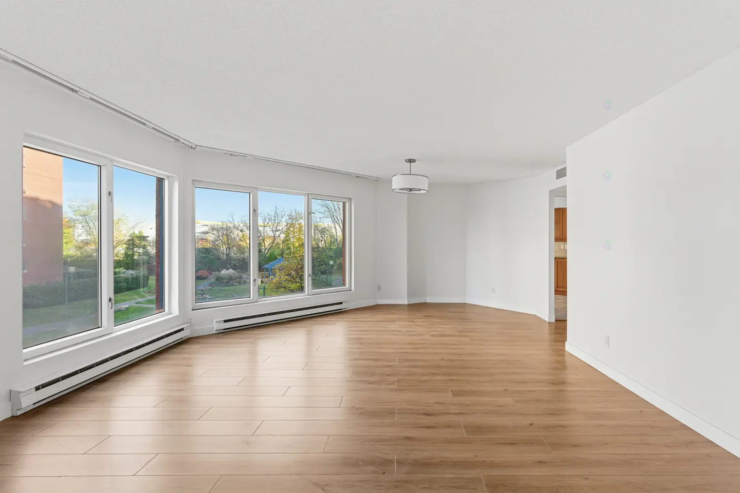 Bright, empty living room with wood floors, white walls, and large windows overlooking a green outdoor space.