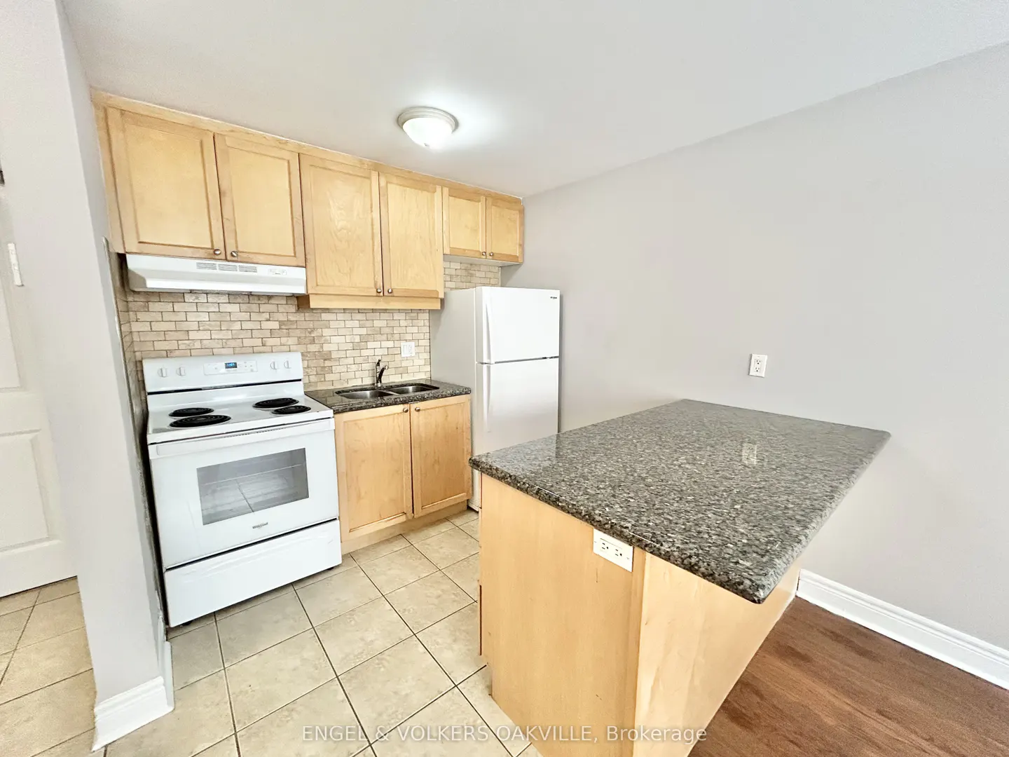 Kitchen with light wood cabinets, white appliances, granite countertops, and a kitchen island on tiled and wood flooring.