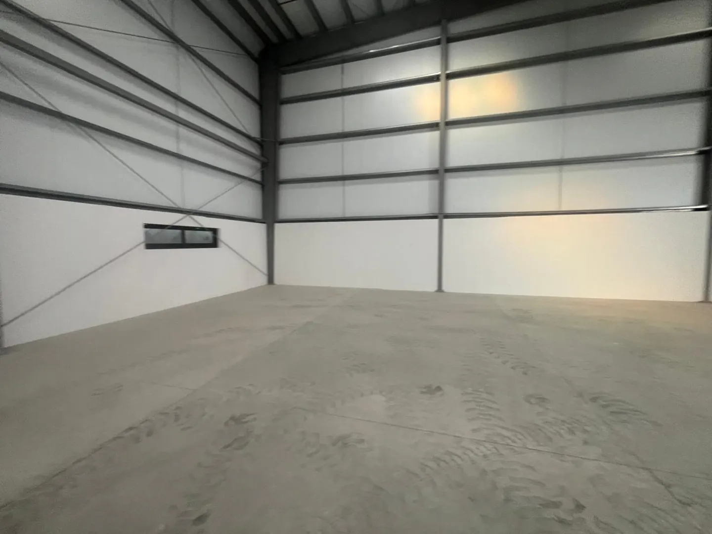An empty warehouse with a concrete floor, white walls, and a metal frame. A small window is visible on the left wall.