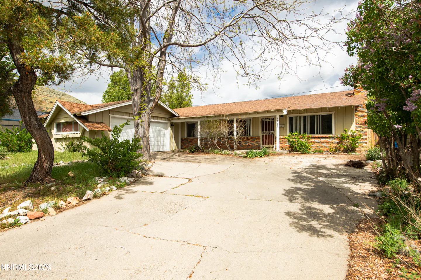 A ranch-style house with a tan exterior, brown roof, and a cracked concrete driveway. Trees frame the house, with mountains visible in the background.