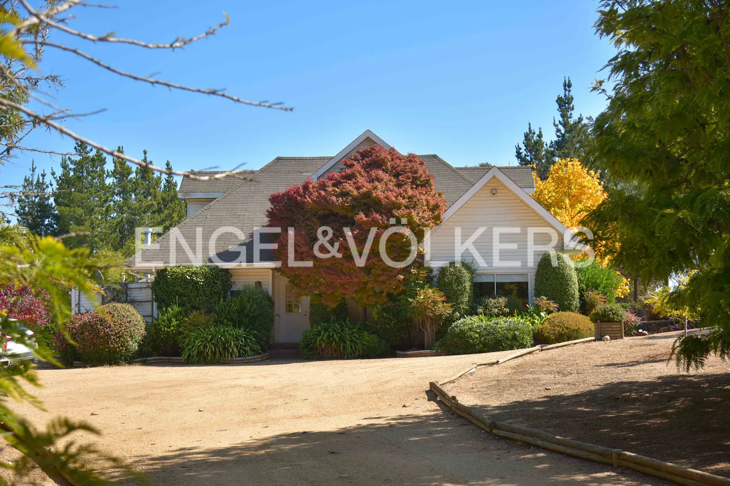 Exterior view of a cream-colored house with a beige roof and a large red tree in front. The Engel & Völkers logo is superimposed on the image.