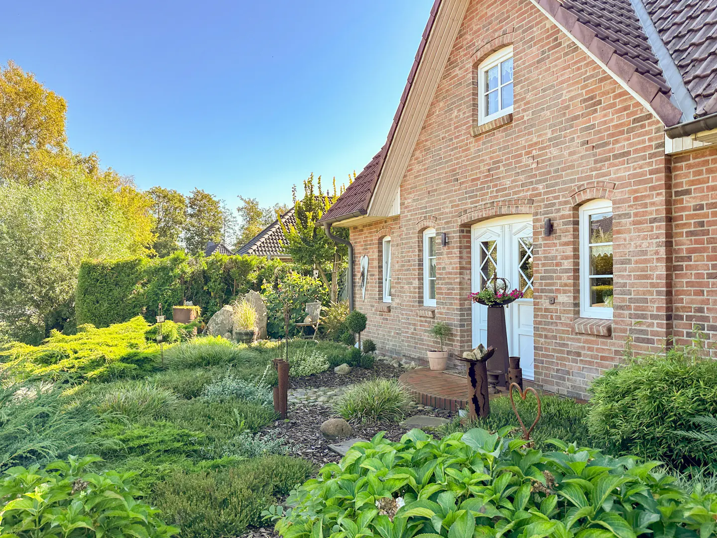 A brick house with white trim and a red roof is surrounded by lush green landscaping. A blue sky is visible above.