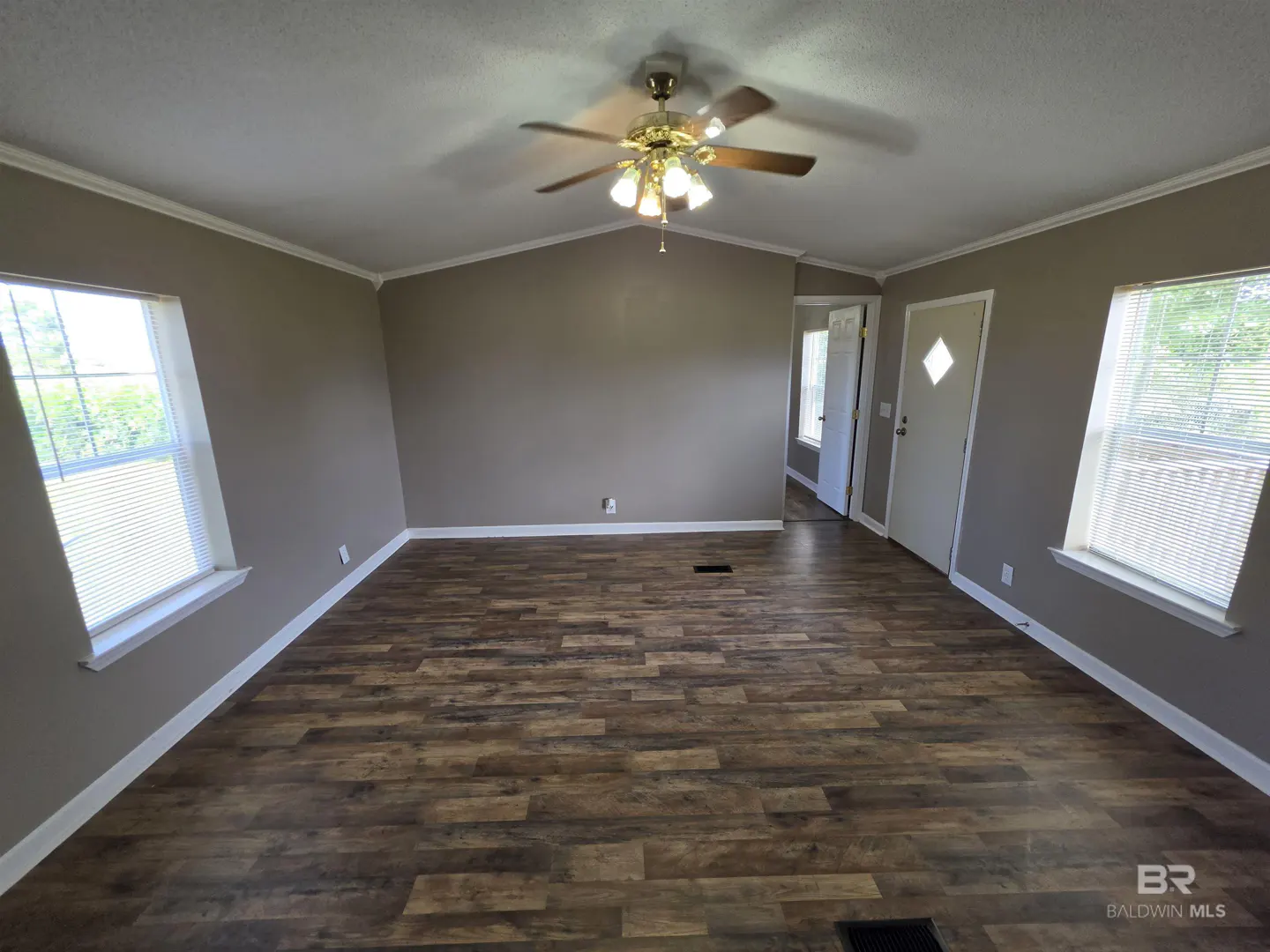 A living room with wood floors, taupe walls, white trim, and a ceiling fan with lights. Two windows with blinds let in natural light. A white door is on the right.