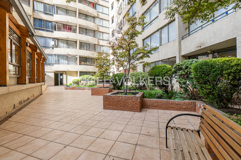 Courtyard view of a multi-story apartment building with trees, bushes, and a wooden bench on a tiled patio.