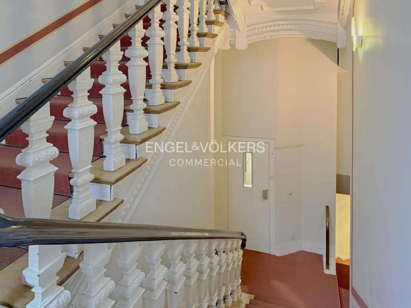 Interior view of a staircase with white balusters, a dark handrail, and red carpeted steps, leading to a white door.
