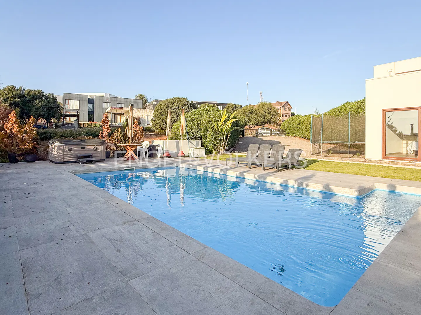 A rectangular swimming pool with blue water, surrounded by a stone patio, lounge chairs, a hot tub, and lush greenery.