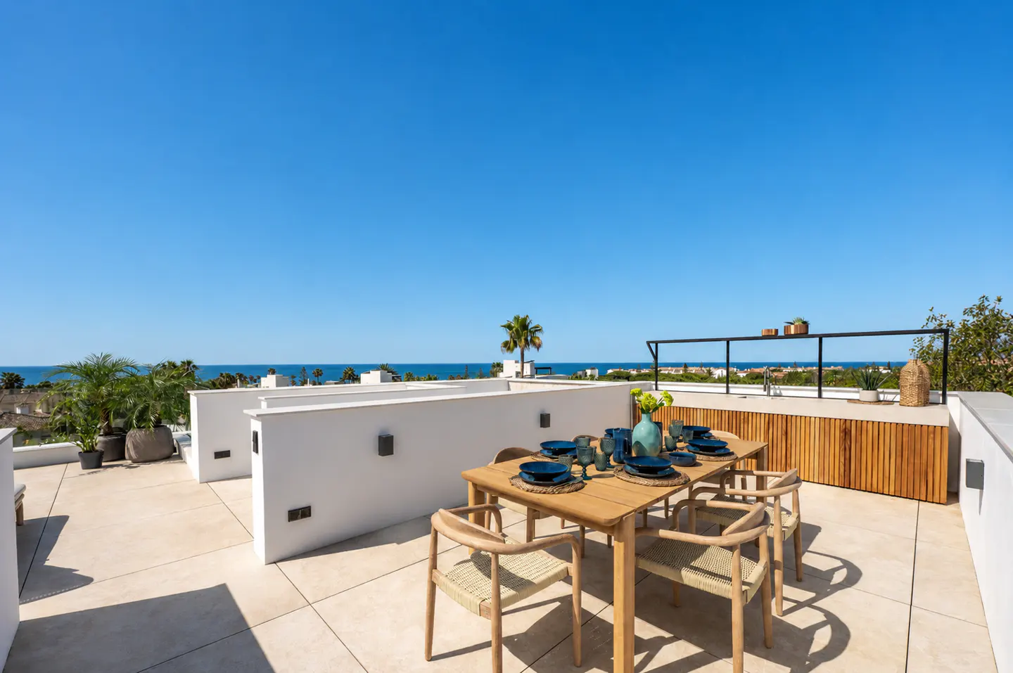 Outdoor dining area on a rooftop terrace with a wooden table, chairs, and blue tableware. Ocean view and clear blue sky in the background.