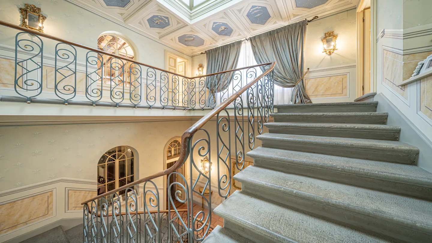 A multi-story foyer with stone stairs, ornate iron railings, and a painted ceiling with a skylight. Pale walls are decorated with sconces and framed panels.