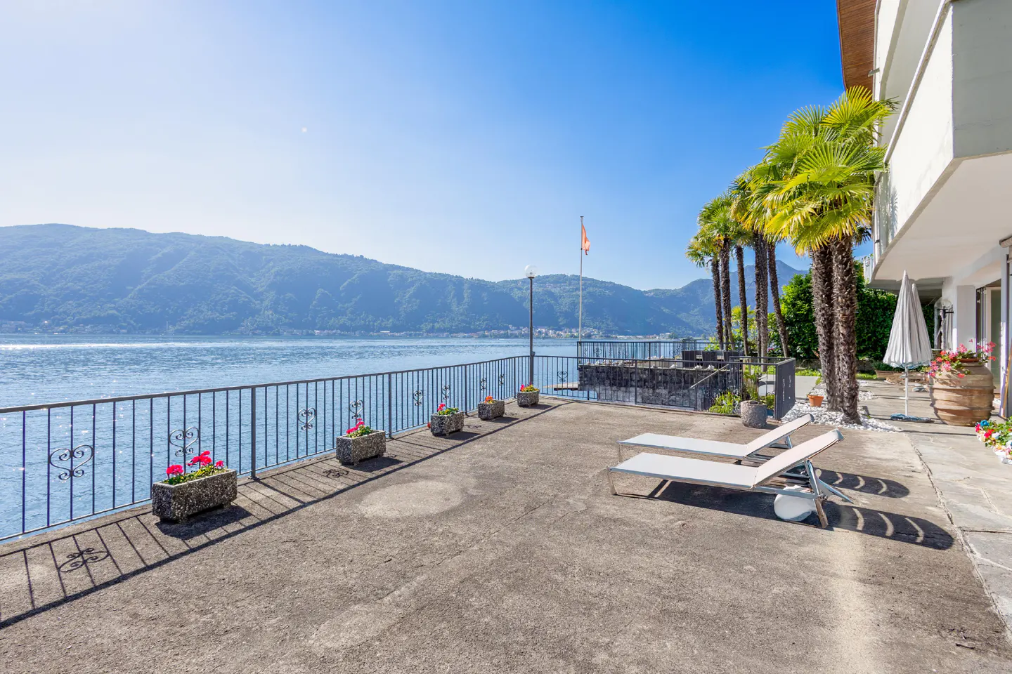 Waterfront patio with lounge chairs, palm trees, and a metal railing overlooking a lake and mountains under a clear blue sky.