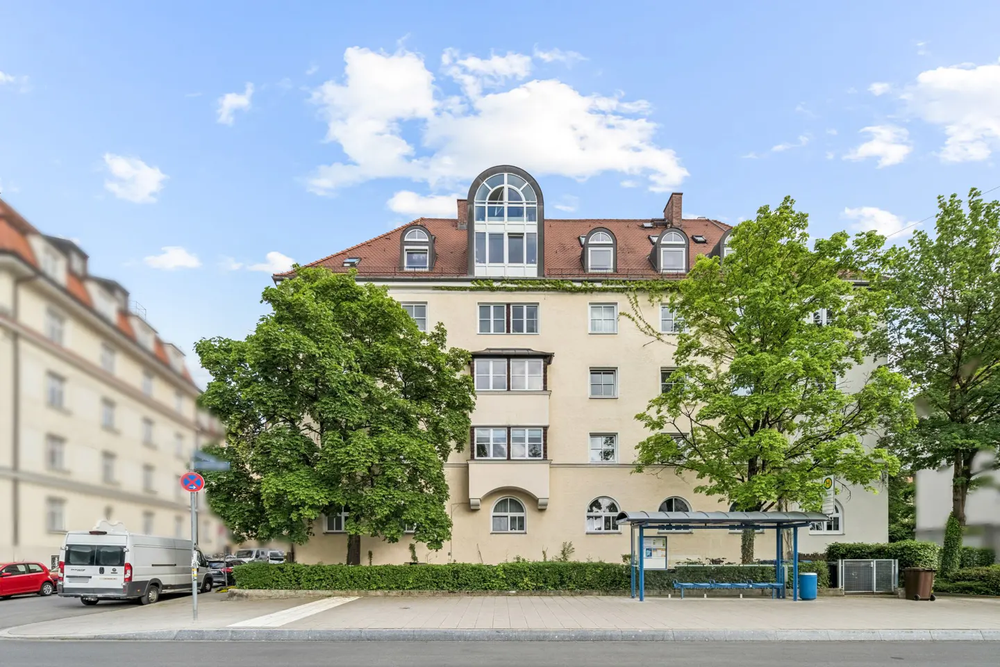 Exterior view of a cream-colored apartment building with a red tile roof and arched windows, framed by green trees and a blue sky.