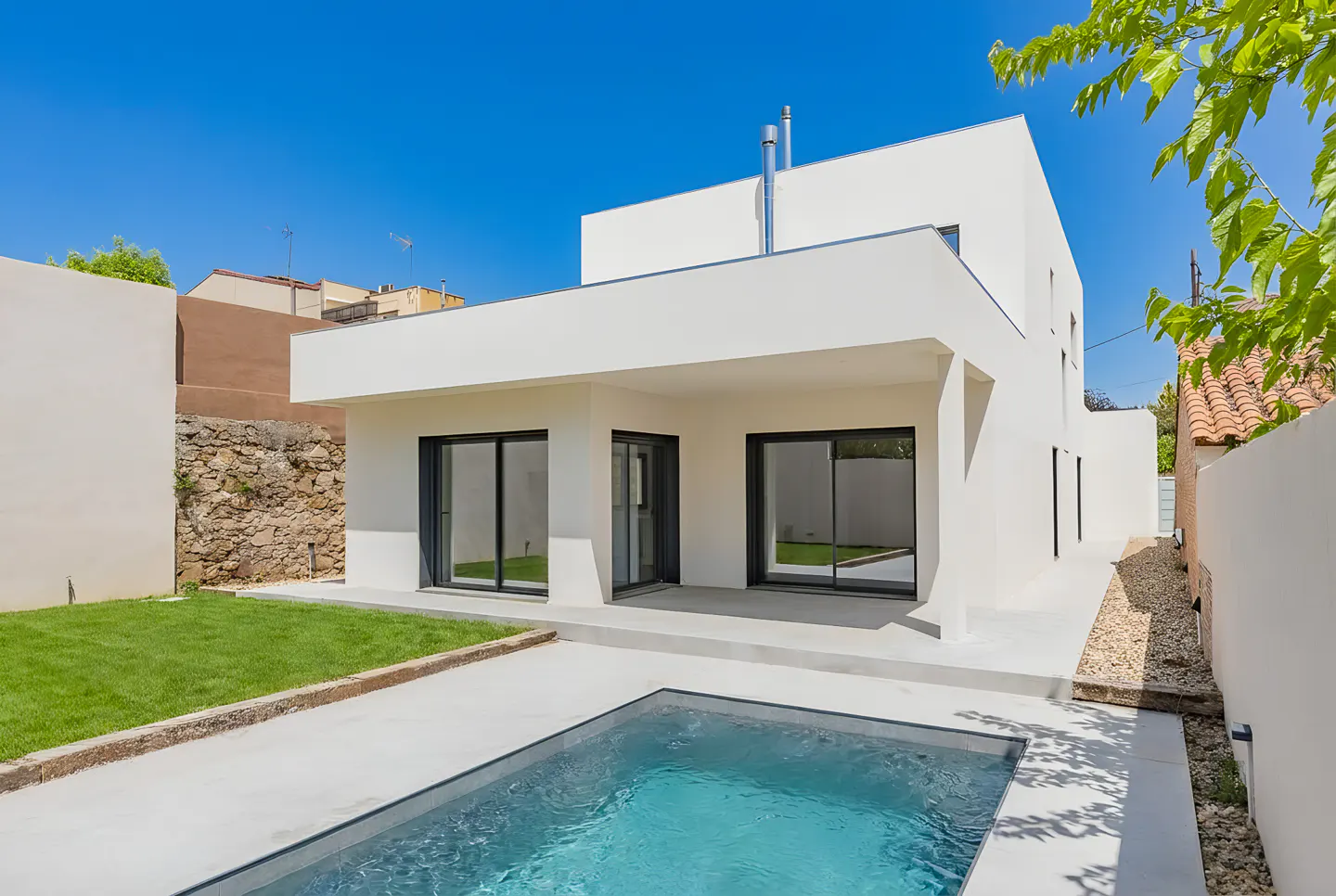 Modern white house with a pool, lawn, and patio under a clear blue sky. Large sliding glass doors offer views into the home.