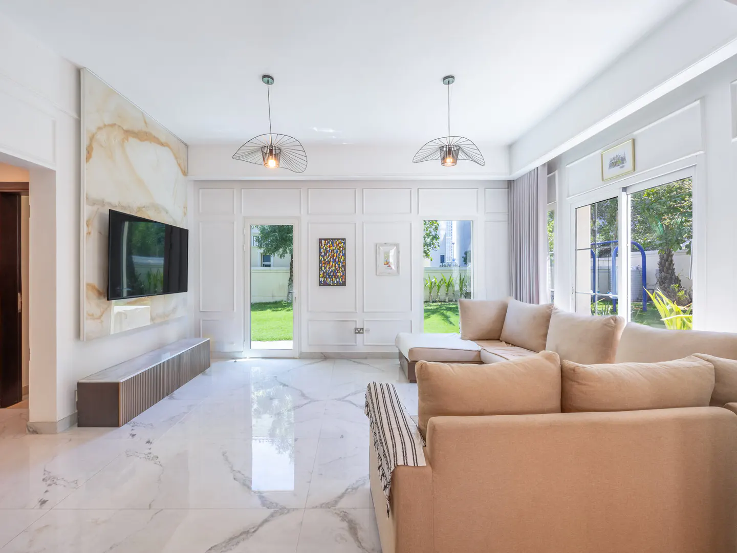 Bright living room with beige sectional sofa, marble floors, and a TV mounted on a marble accent wall. Doors open to a green lawn.