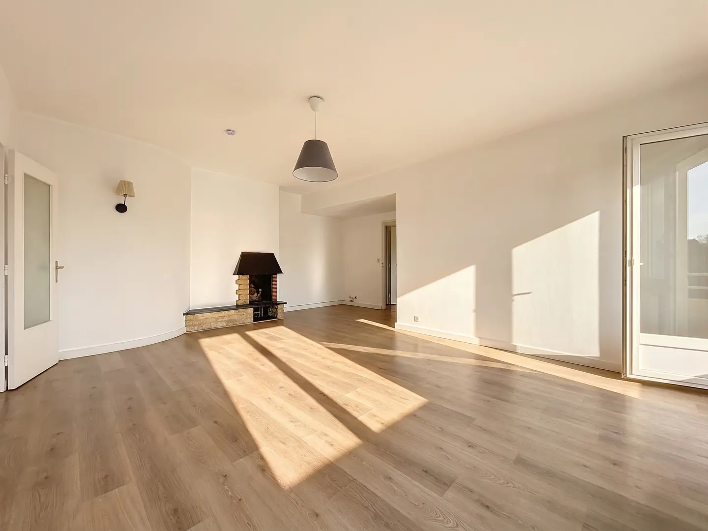 Bright, empty room with wood floors, white walls, and a stone fireplace. Sunlight streams in from a glass door.
