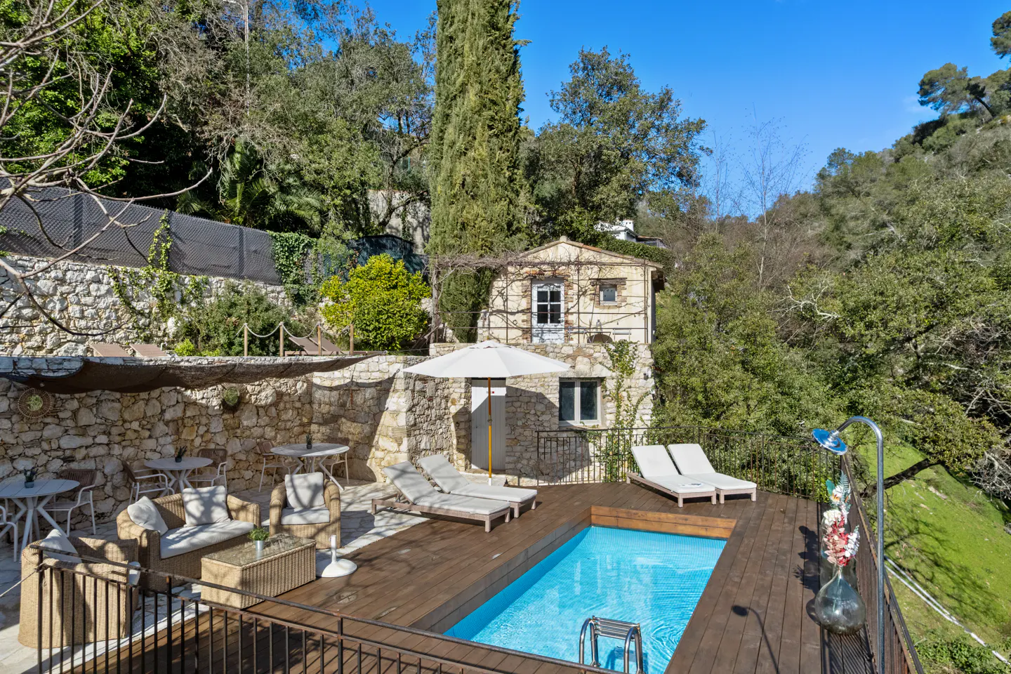 Outdoor pool area with lounge chairs, tables, and seating on a wooden deck. Stone building and lush greenery in the background.