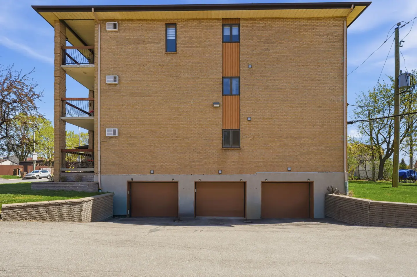 Tan brick apartment building with three brown garage doors. Balconies on the left side and windows on the right. Blue sky background.