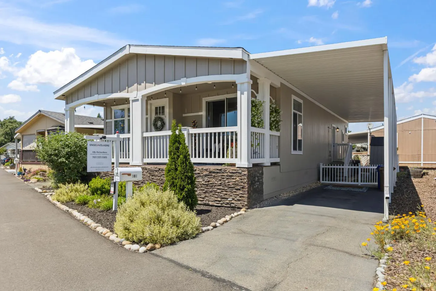 Tan single-story home with white trim, a covered porch, and a carport on a sunny day. A "For Sale" sign sits in the landscaped yard.