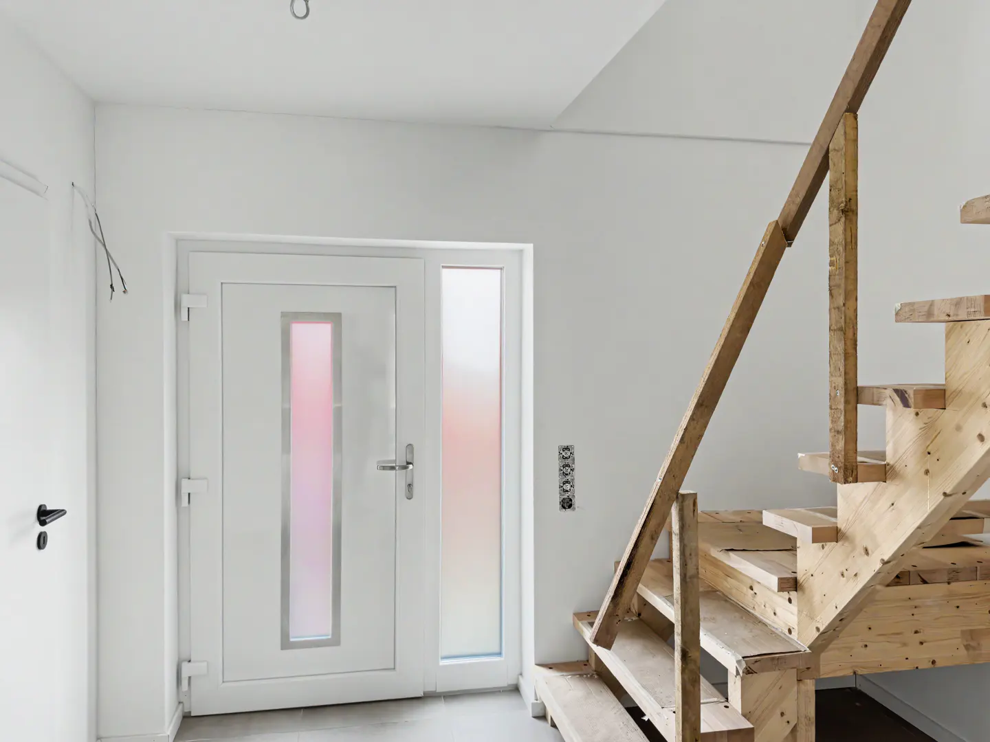 Modern white front door with frosted glass panels next to an unfinished wooden staircase in a bright, minimalist entryway.