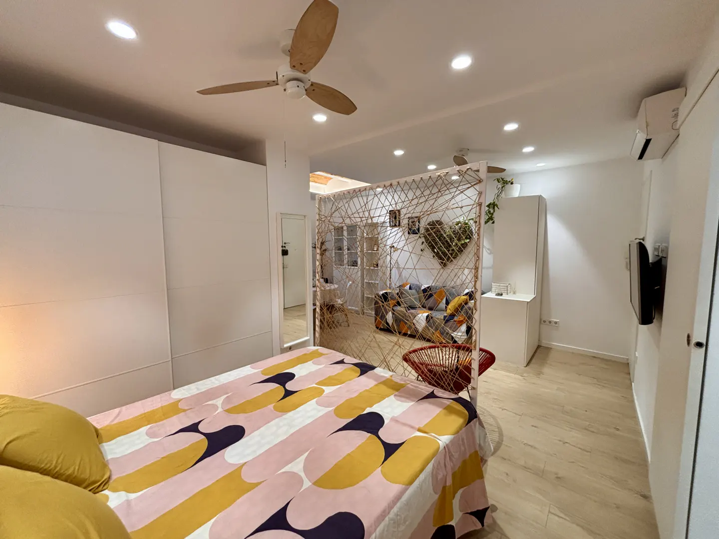 Modern bedroom with a patterned bedspread, a rope room divider, and a cozy seating area with a heart-shaped wall decoration.