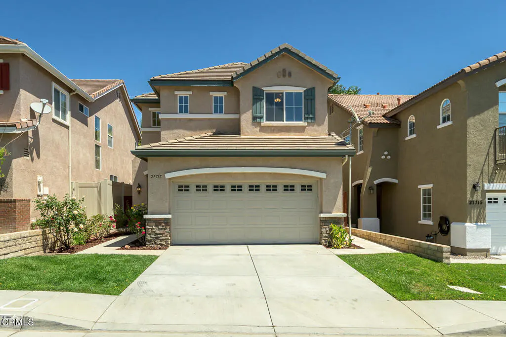 Two-story beige house with a large garage door and green trim under a clear blue sky.