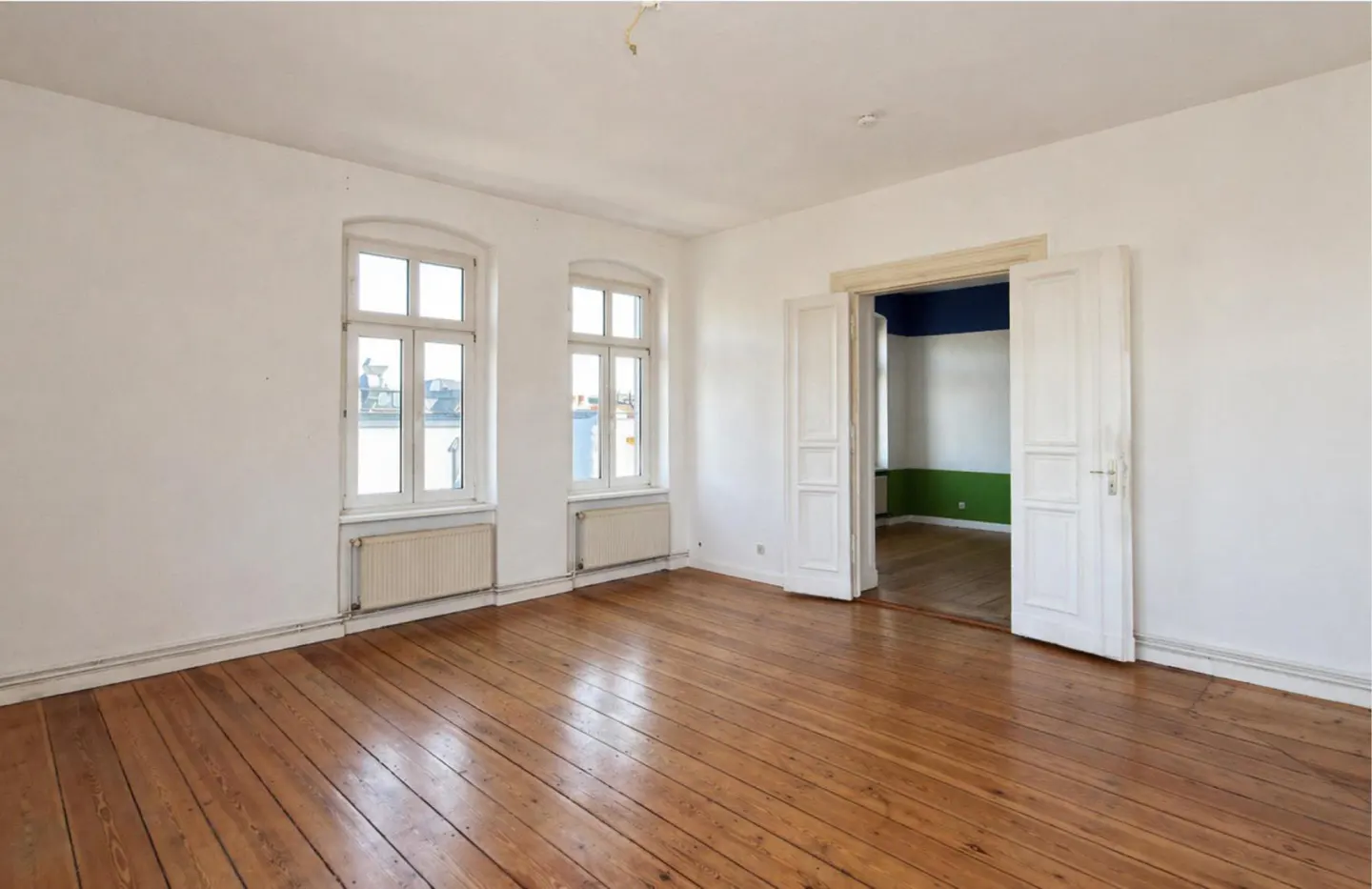 Bright, empty room with wood floors, white walls, and two windows. An open doorway leads to a room with blue and green walls.
