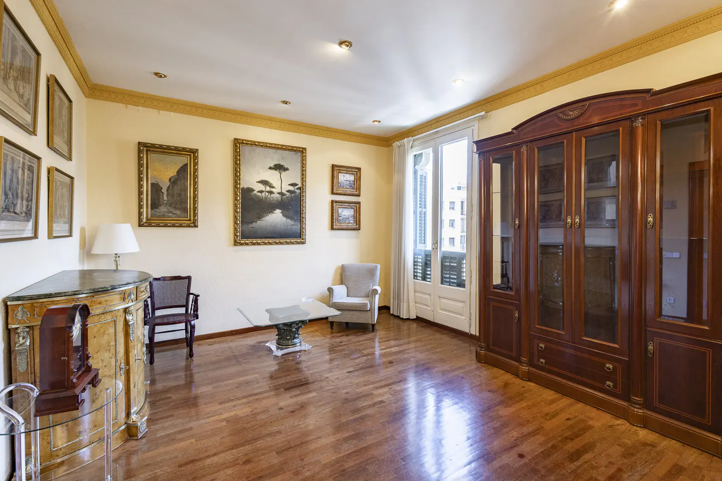 A living room with hardwood floors, cream walls, and gold trim. There is a large wooden cabinet, paintings on the wall, and a glass coffee table.