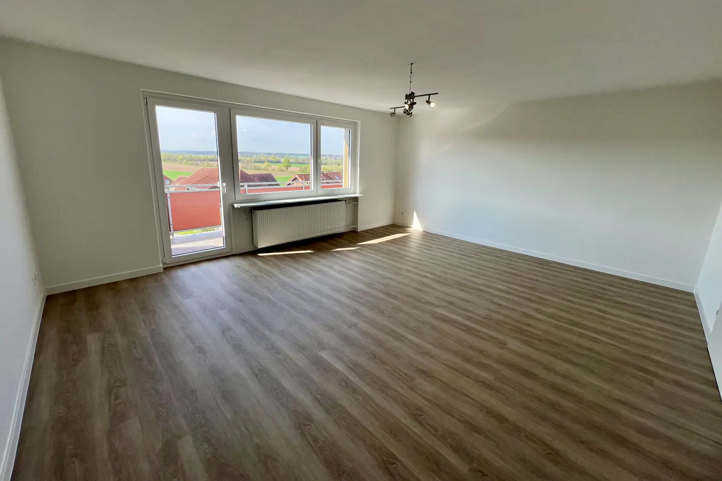 Bright, empty room with wood-look flooring, white walls, and a window with a view. A radiator sits beneath the window. A modern light fixture hangs from the ceiling.