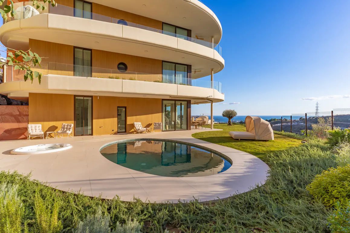 A modern, multi-story tan house with a curved design, a pool, and a green lawn under a blue sky.