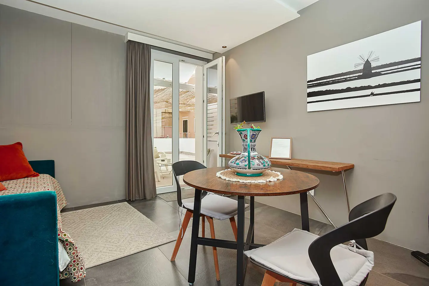 Cozy apartment interior with a round wooden table, black chairs, and a blue sofa with a red pillow. Balcony doors open to an outdoor seating area.