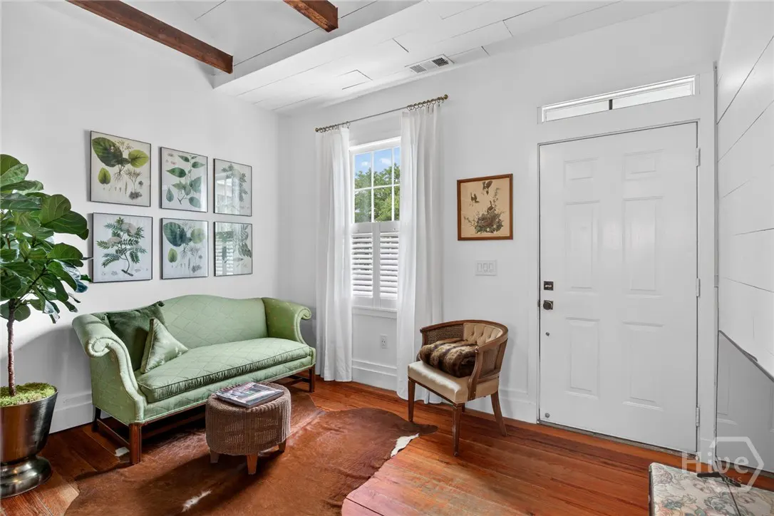 Living room with a green sofa, wooden floors, and white walls. Botanical prints hang above the sofa. A white front door is on the right.