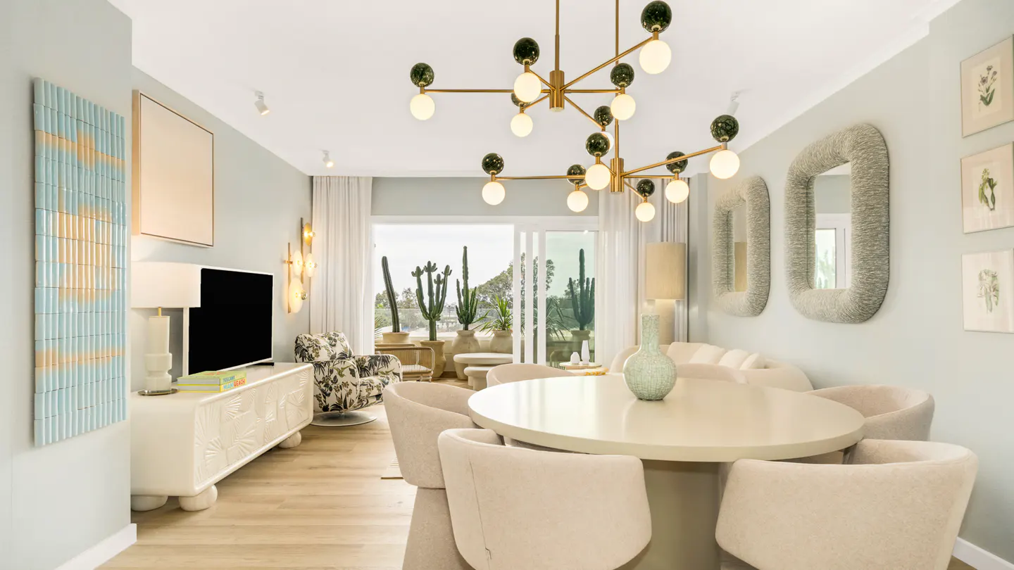 Bright, modern dining room with a round table, cream chairs, and a gold chandelier. Cacti are visible through the open patio doors.
