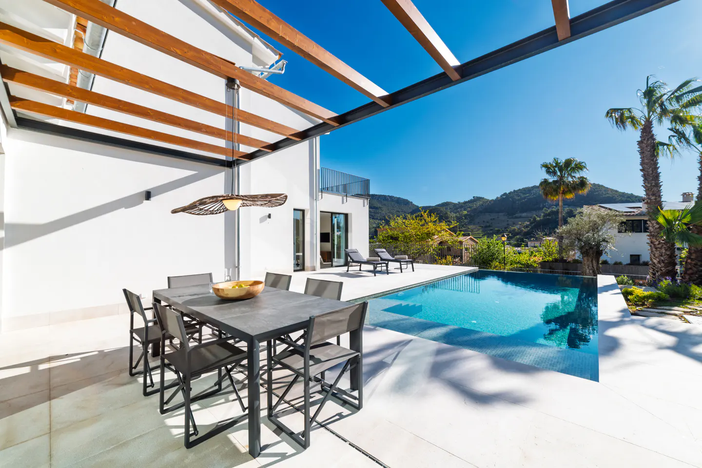 Outdoor patio with a dining table, chairs, and a pool. Mountains and palm trees are in the background.