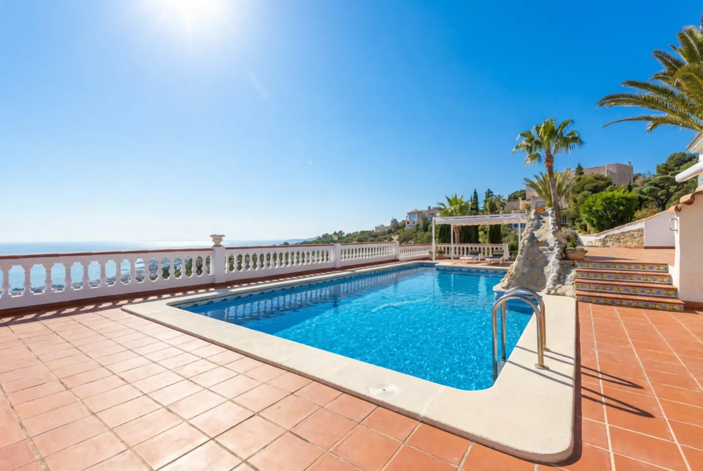 Outdoor pool with blue water and metal ladder, surrounded by terracotta tiles. Ocean view with white railing and palm trees under a clear blue sky.