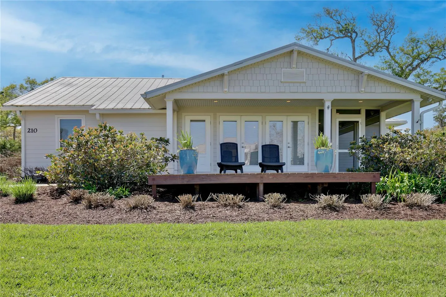 A single-story house with a metal roof, a wooden porch with two chairs, and green grass.