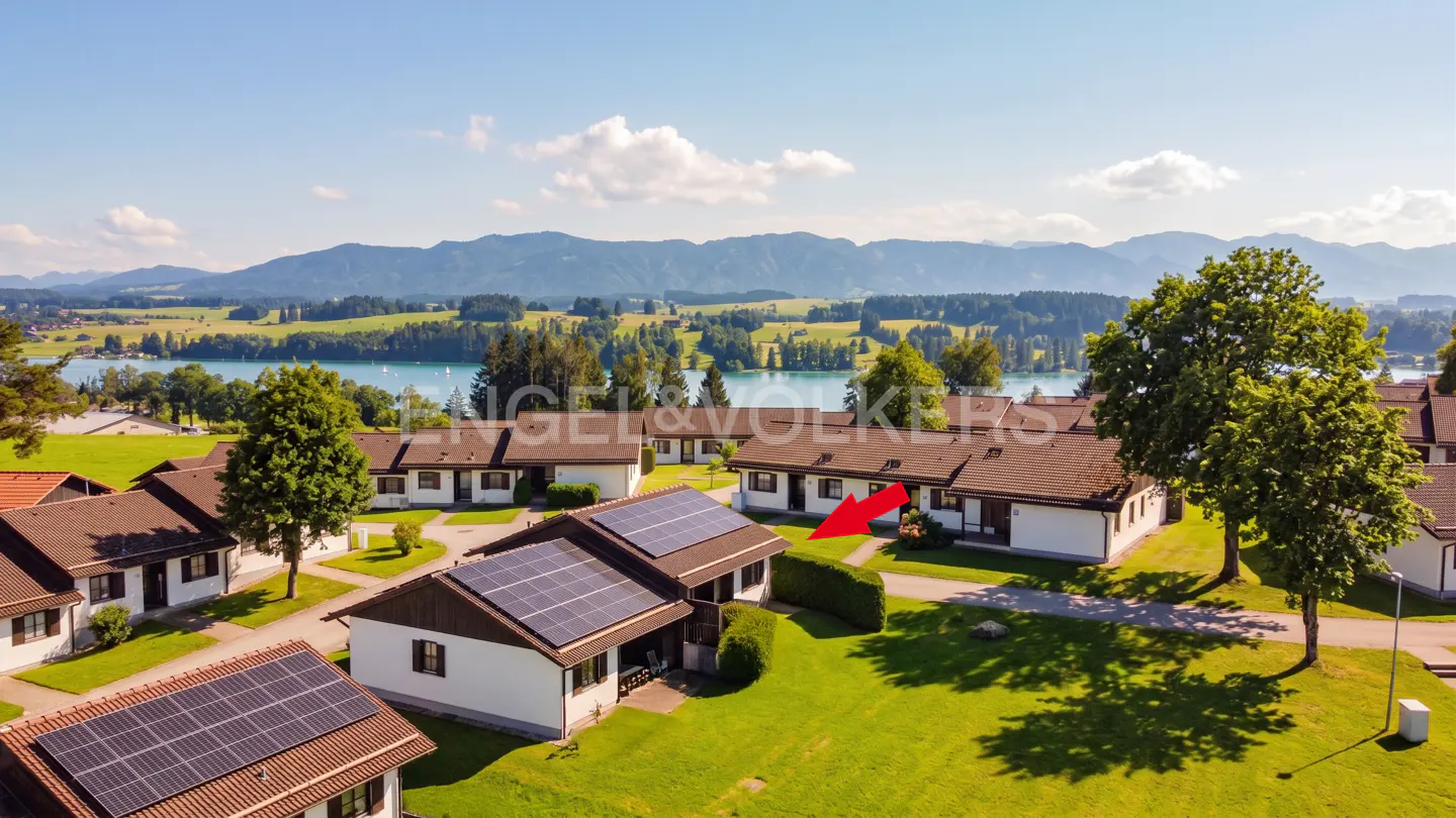 Aerial view of white houses with solar panels on roofs, green lawns, and a lake with mountains in the background. Red arrow points to a house.